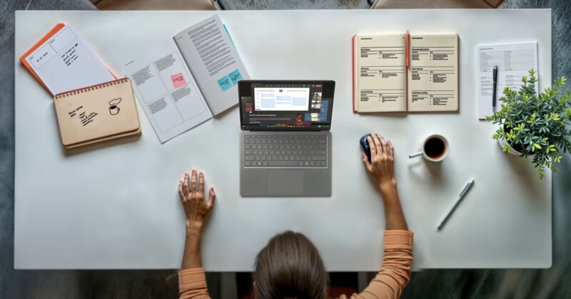 A person sits at a tidy desk with a laptop, notebooks, papers, a pen, a coffee cup, and a small potted plant, working with one hand on the mouse and the other resting on the desk.